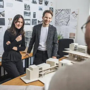 Zwei Architekten, ein Mann und eine Frau, stehen lächelnd in einem hellen Büro vor Architekturmodellen und einer Wand mit Entwürfen – Businessfotografie aus Ulm.