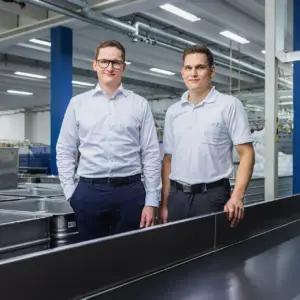 Zwei lächelnde Männer in professioneller Arbeitskleidung (Hemd und Poloshirt) in einer modernen Industrieumgebung – Businessfotografie aus Ulm.