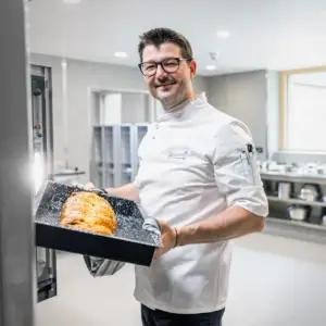 Ein lächelnder Koch in weißer Arbeitskleidung präsentiert ein frisch gebackenes Tablett in einer modernen Edelstahl-Großküche – Gastronomiefotografie aus Ulm.