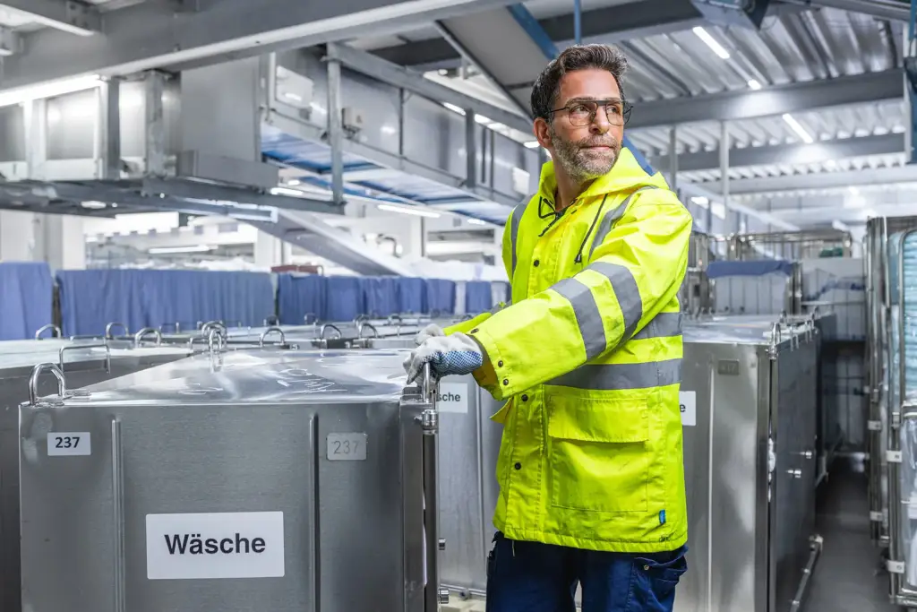 Logistik-Mitarbeiter in neongelber Warnschutzjacke bewegt Transportcontainer in einer Ulmer Werkshalle.