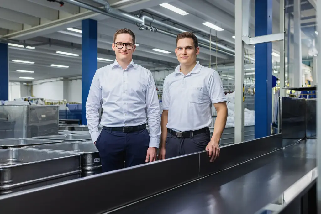 Zwei junge männliche Mitarbeiter stehen gemeinsam in einem modernen Ulmer Unternehmensraum für ein Teamfoto.