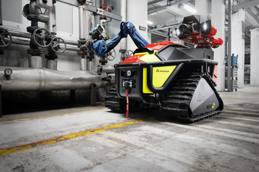 Der rote Löschroboter Magirus Wolf R1 bedient mit seinem blauen Roboterarm ein Ventil an einer industriellen Rohrleitungsanlage in einer hellen Werkshalle.