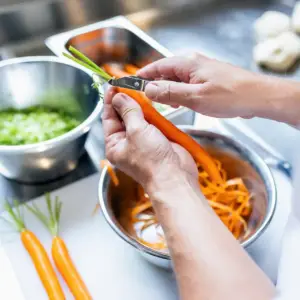 Nahaufnahme der Hände eines Kochs beim Schälen einer frischen Karotte in einer Edelstahl-Küche – professionelle Werbefotografie aus Ulm.